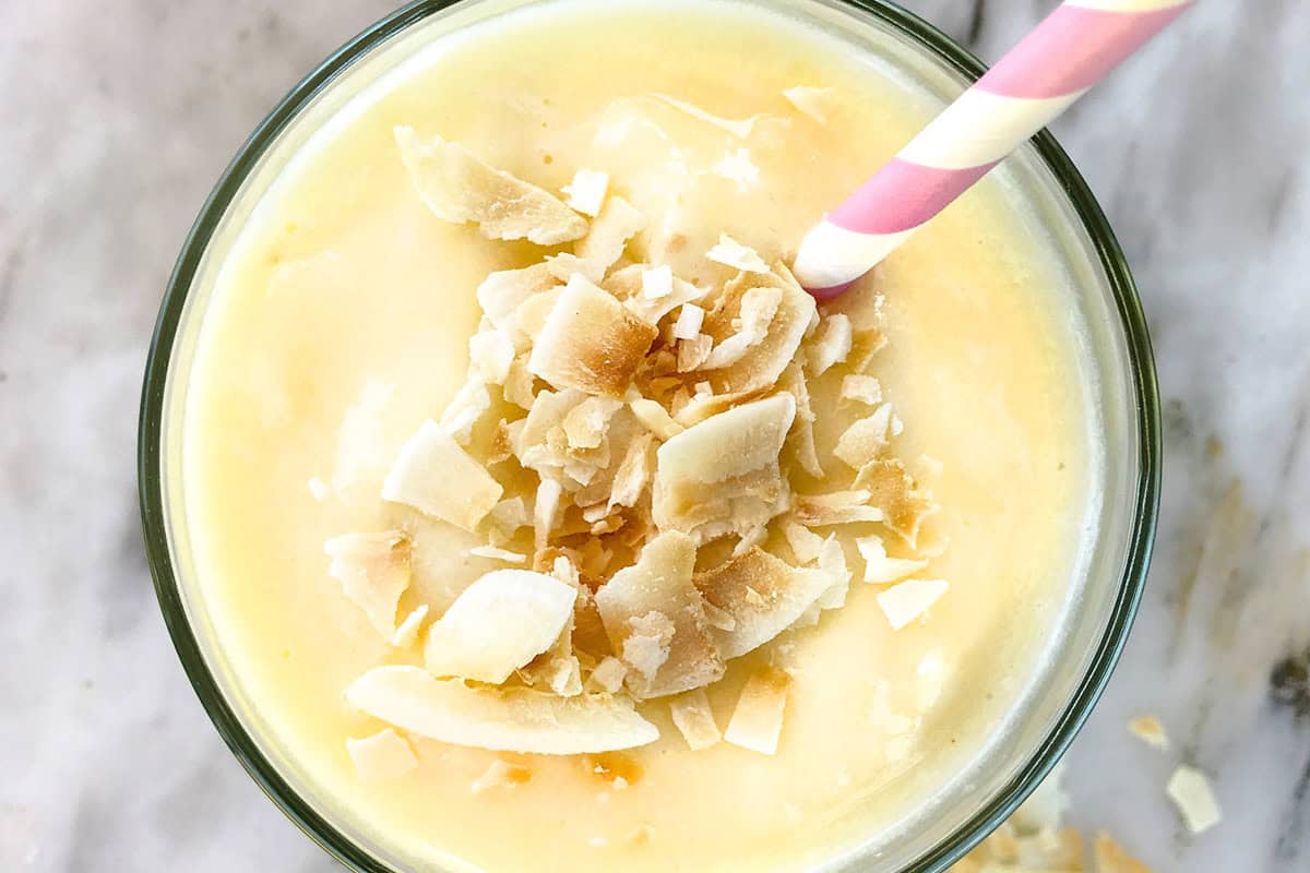 A creamy yellow pineapple smoothie topped with toasted coconut flakes, served in a glass with a pink-and-white striped straw on a light, marble surface.