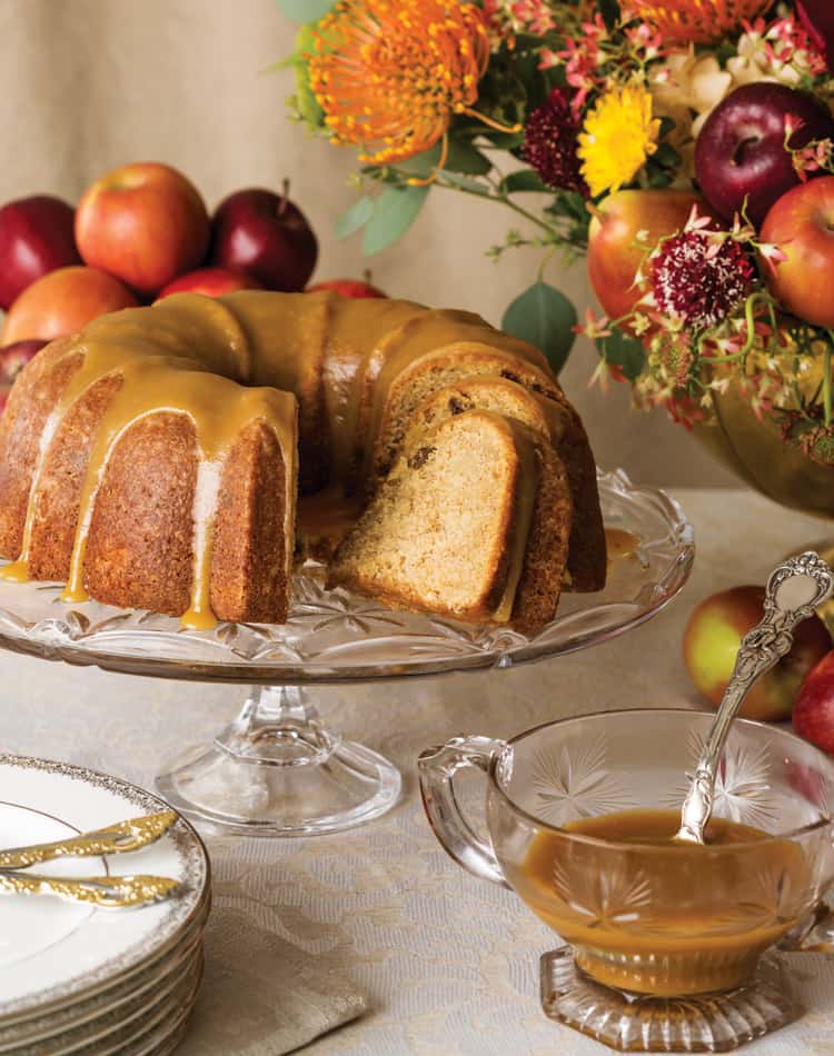 Bundt cake with caramel glaze on a glass stand, surrounded by apples and fall flowers-perfect for an Autumn Afternoon Tea-served with a glass sauce boat of caramel sauce on a table set with plates and utensils.