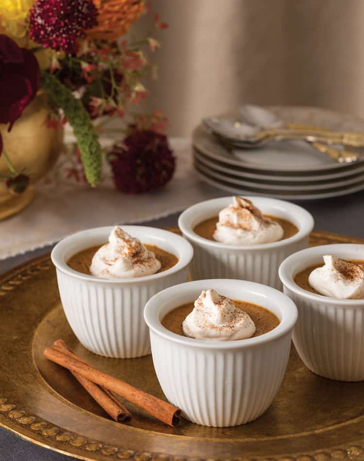 Four white ramekins filled with dessert, topped with whipped cream and cinnamon, sit on a gold tray-perfect for an Autumn Afternoon Tea-while cinnamon sticks and a floral arrangement add warmth in the background.