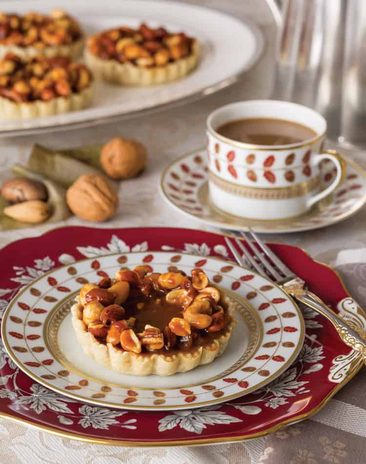 A nut tart sits on a decorative plate next to a fork and knife, with a cup of coffee and whole nuts in the background-perfect for an Autumn Afternoon Tea.