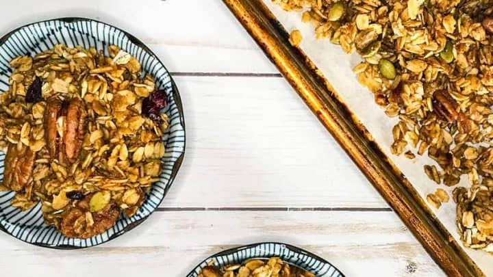 Two small bowls of pumpkin spice granola with nuts and dried fruit are placed on a white wooden surface next to a baking sheet lined with fresh granola.