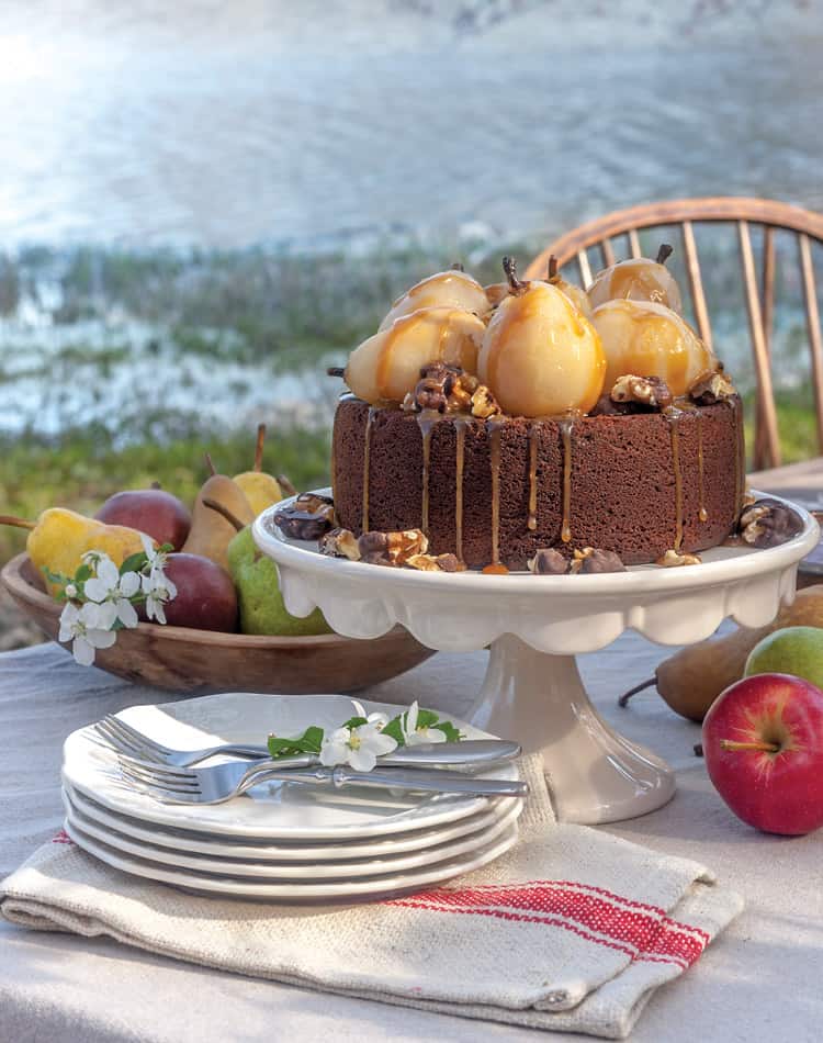A chocolate cake topped with whole poached pears and caramel drizzle sits on a cake stand on an outdoor table, surrounded by apples, pears, plates, and utensils-perfect for an Autumn Afternoon Tea.