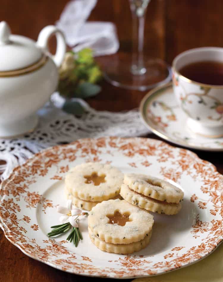 Three powdered sugar sandwich cookies with a flower-shaped cutout rest on a decorative plate, next to a teacup, teapot, lace napkin, and small flower garnish-perfect for an Autumn Afternoon Tea.