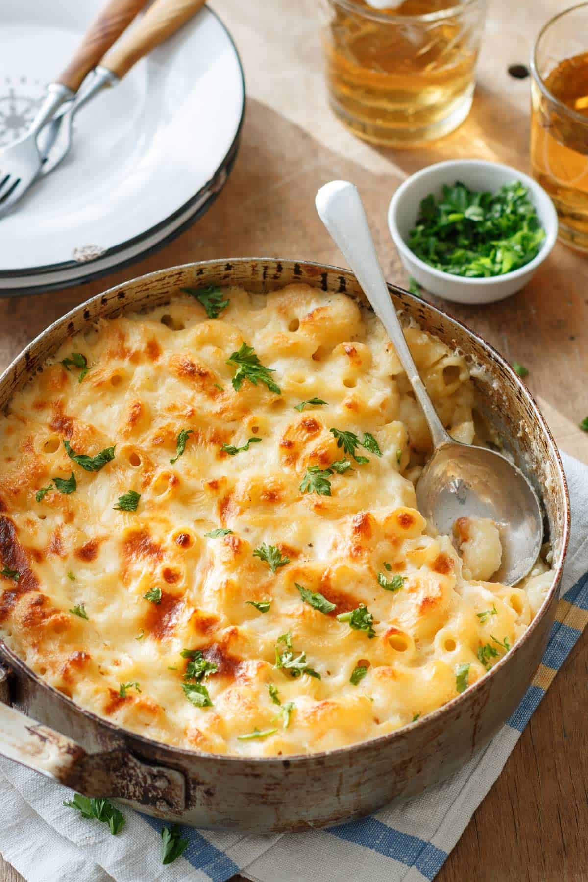 A delectable baked mac and cheese dish in a round pan, topped with golden melted cheese and garnished with parsley. A serving spoon rests in the creamy delight. Nearby are plates, forks, a small bowl of parsley, and two glasses of iced tea on a wooden table.