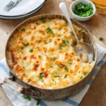 A classic Baked Mac and Cheese dish sits in a round metal pan, topped with perfectly melted cheese and a sprinkle of fresh parsley, with a serving spoon resting inside. Nearby, a small bowl of chopped herbs adds an extra touch of flavor to this comforting favorite.