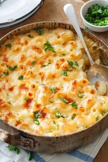 A classic Baked Mac and Cheese dish sits in a round metal pan, topped with perfectly melted cheese and a sprinkle of fresh parsley, with a serving spoon resting inside. Nearby, a small bowl of chopped herbs adds an extra touch of flavor to this comforting favorite.