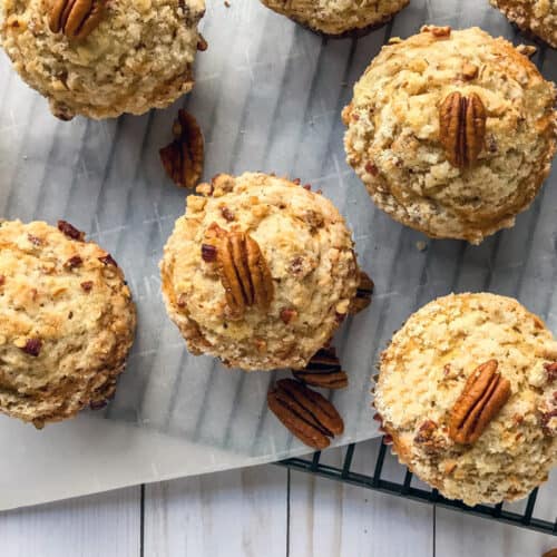Freshly baked maple pecan muffins topped with pecan halves are arranged on a piece of parchment paper over a cooling rack. Scattered pecan pieces are visible around the muffins.