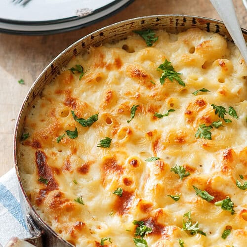 A delicious baked mac and cheese dish is topped with golden brown melted cheese and garnished with fresh parsley. A silver serving spoon rests inside, inviting you to serve up creamy goodness. A small bowl of chopped parsley waits in the background for added flavor.