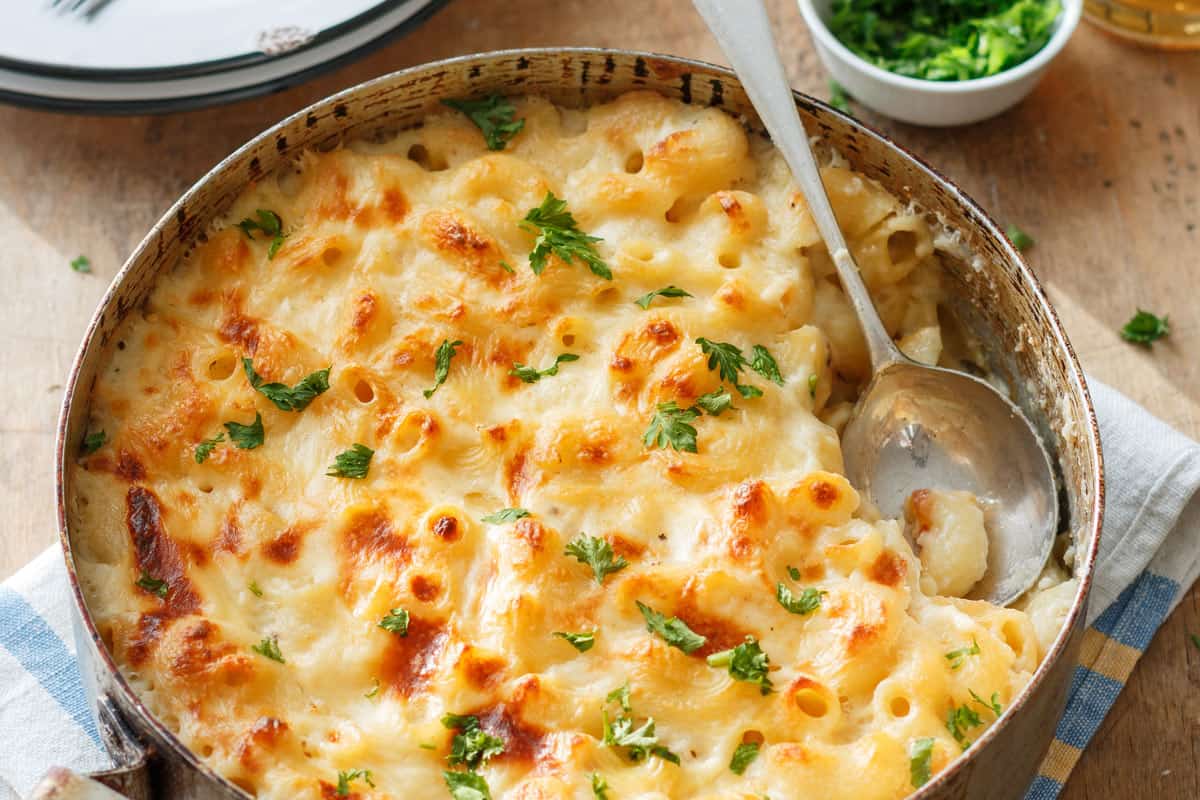 A creamy baked mac and cheese dish topped with golden-brown cheese and garnished with fresh parsley, served in a round pan with a spoon on a wooden table. A small bowl of chopped herbs is in the background.