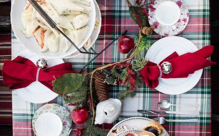 Overhead view of a festive Christmas Tea table set with assorted desserts, sandwiches, teacups, a teapot, red napkins, and holiday decorations on a plaid tablecloth.