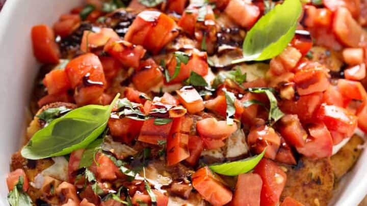 A white dish filled with breaded eggplant slices topped with diced tomatoes, mozzarella cheese, fresh basil leaves, and a drizzle of balsamic glaze, reminiscent of classic bruschetta chicken flavors.