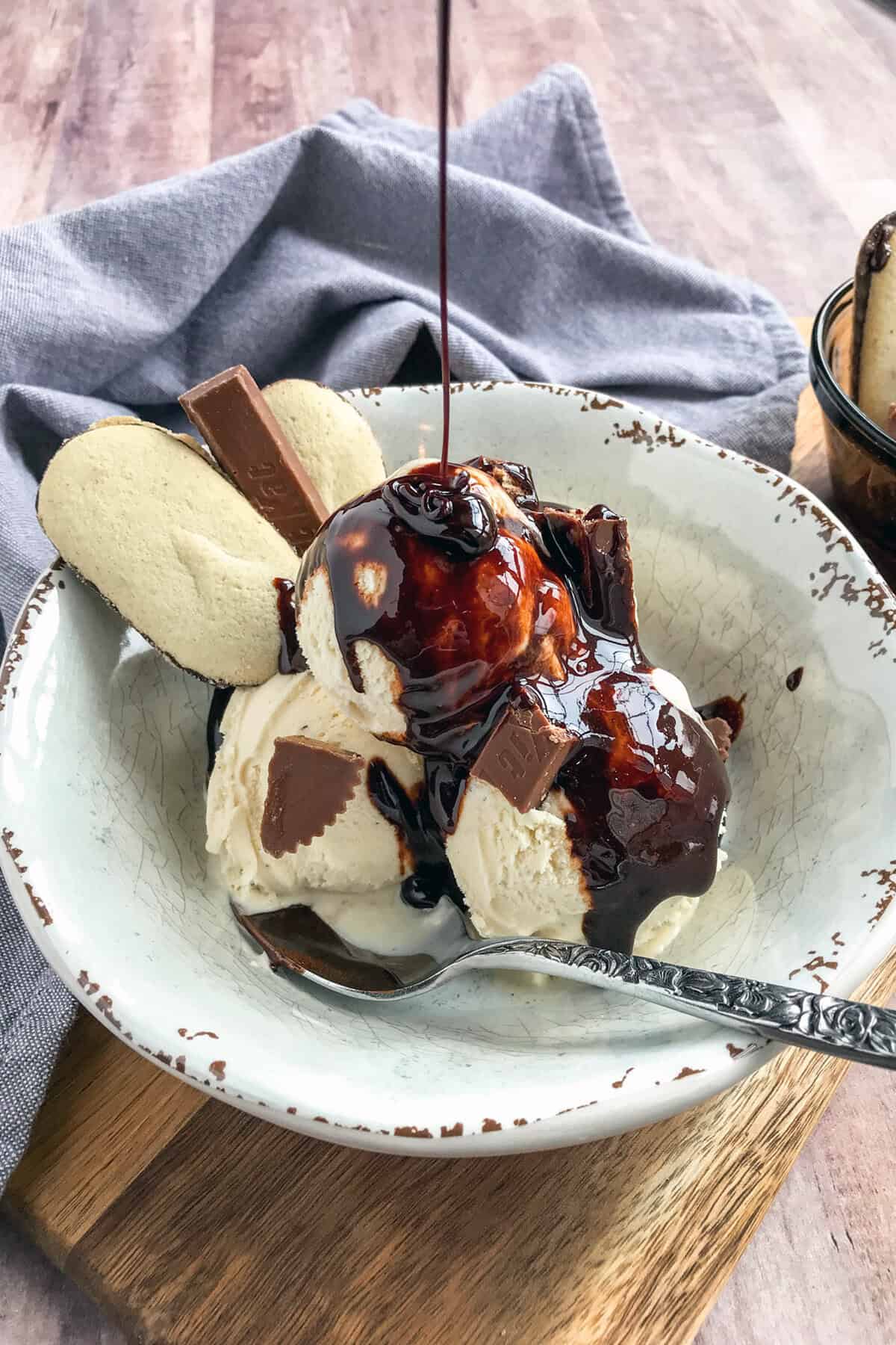 A bowl of vanilla ice cream topped with chocolate sauce and chocolate pieces, garnished with two cookies. A spoon rests in the bowl, and a gray cloth napkin is in the background on a wooden surface.