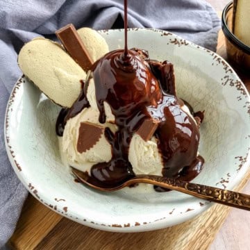 A bowl of vanilla ice cream topped with rich chocolate sauce, chocolate pieces, and two cookies, with a spoon resting in the bowl.