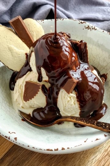A bowl of vanilla ice cream topped with rich chocolate sauce, chocolate pieces, and two cookies, with a spoon resting in the bowl.