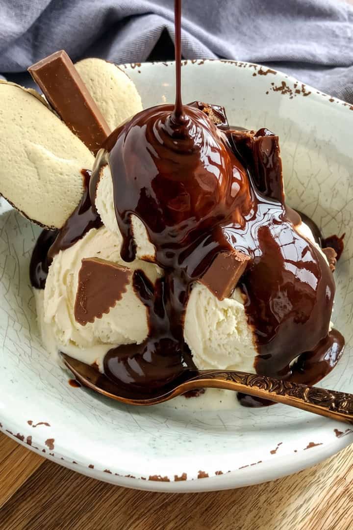 A bowl of vanilla ice cream topped with rich chocolate sauce, chocolate pieces, and two cookies, with a spoon resting in the bowl.