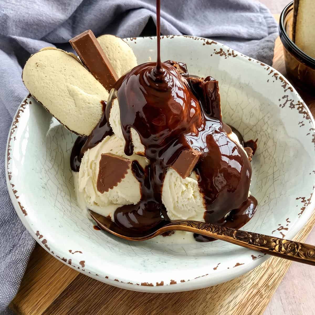 A bowl of vanilla ice cream topped with rich chocolate sauce, chocolate pieces, and two cookies, with a spoon resting in the bowl.