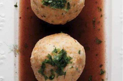 Three round bread dumplings, a classic German Christmas food, are topped with chopped herbs and arranged on a rectangular plate with dark brown sauce, next to a silver spoon.