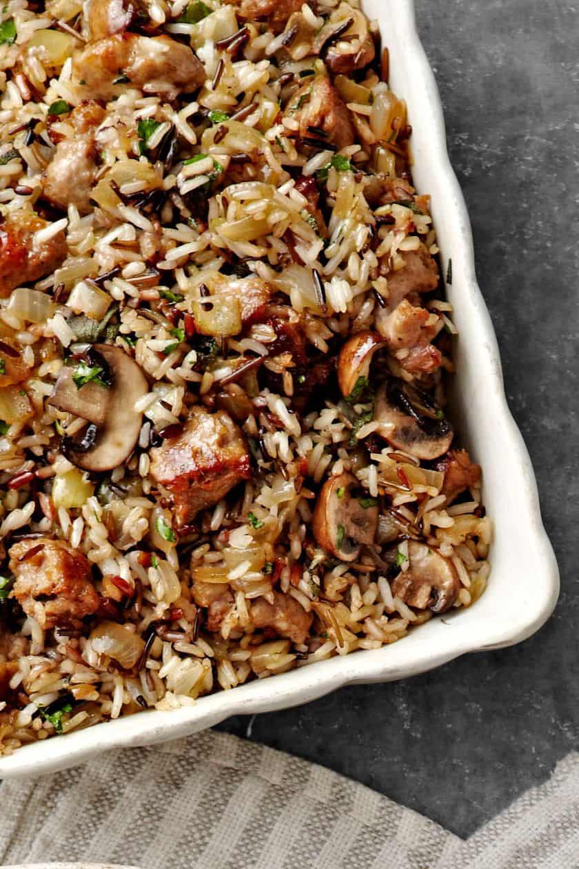 A close-up of a casserole dish filled with wild rice, sausage, mushrooms, and herbs-perfect inspiration for Thanksgiving stuffing recipes-placed on a gray surface with a striped cloth nearby.
