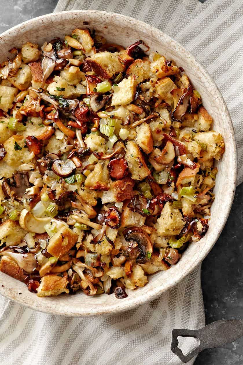 A bowl of stuffing with chunks of bread, mushrooms, celery, onions, and herbs sits on a striped cloth-perfect inspiration for delicious Thanksgiving stuffing recipes.