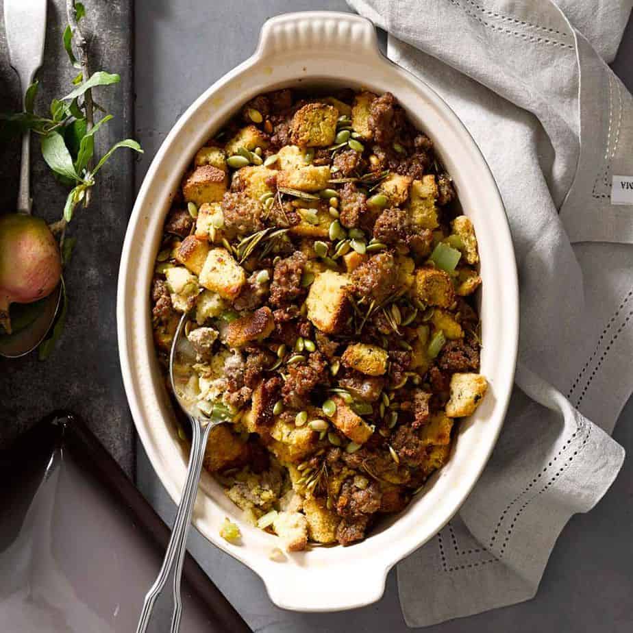 Oval dish filled with bread stuffing mixed with sausage, herbs, and seeds-a savory twist on classic Thanksgiving stuffing recipes-served with a large spoon on a gray surface beside a napkin.