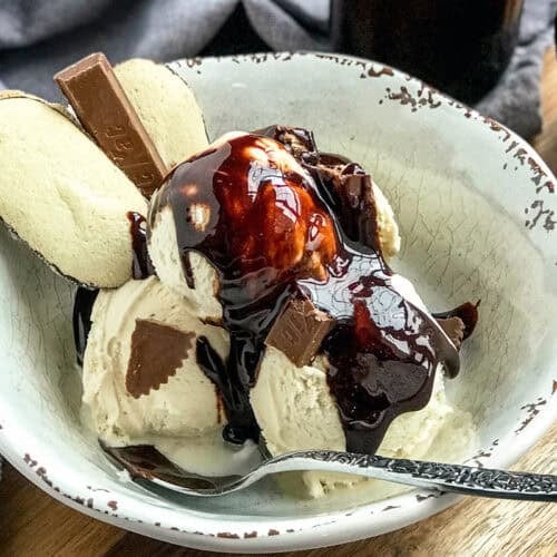 A bowl of vanilla ice cream topped with rich chocolate sauce, broken chocolate pieces, and two wafer cookies, with a spoon resting in the bowl.