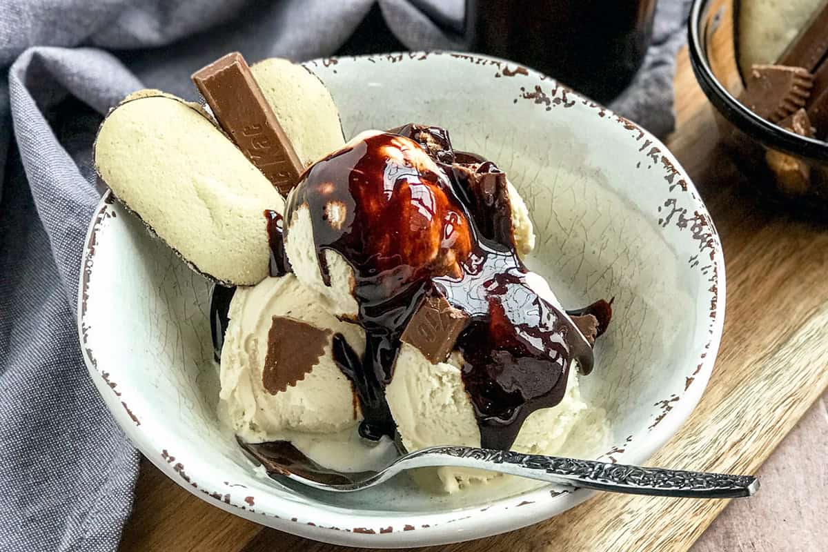 A bowl of vanilla ice cream topped with rich chocolate sauce, broken chocolate pieces, and two wafer cookies, with a spoon resting in the bowl.