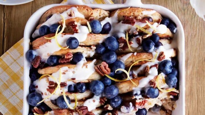 A baking dish filled with cinnamon rolls topped with blueberries, pecans, icing, and lemon zest—perfect for Make Ahead Breakfast Casseroles—sits on a wooden table beside a yellow-checked napkin and a vase with white tulips.