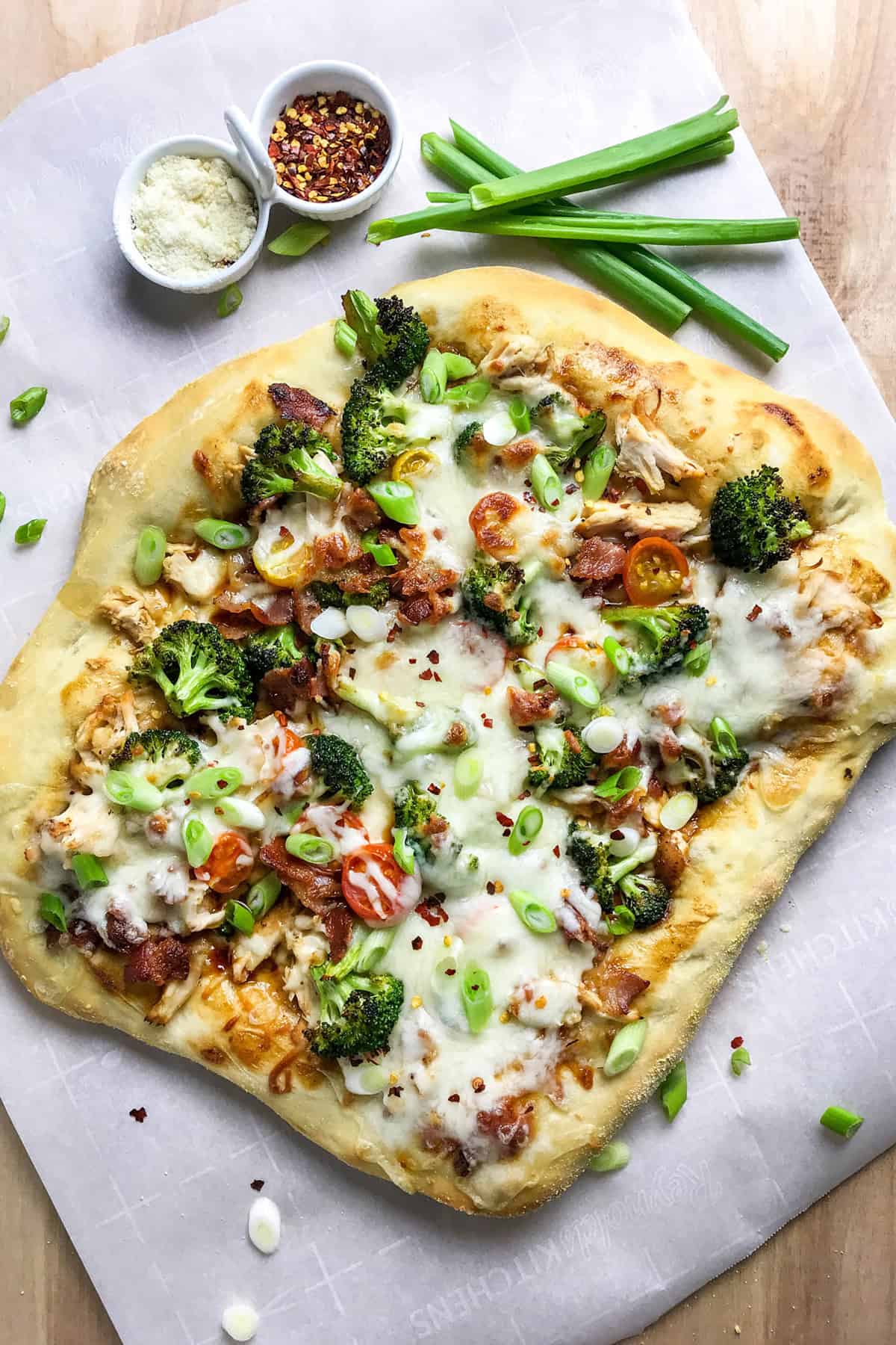 Top view of homemade teriyaki chicken pizza on a parchment lined wooden surface with green onions and red pepper flakes for serving.