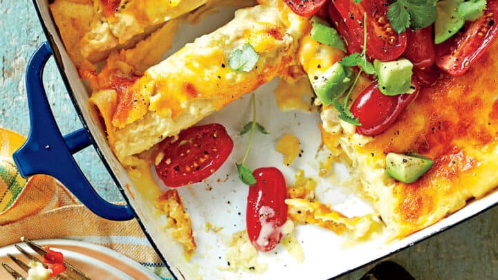 A baked cheesy casserole, perfect for Make Ahead Breakfast Casseroles, with a piece cut out, topped with halved cherry tomatoes, avocado chunks, and fresh herbs; a serving is plated nearby with a fork.