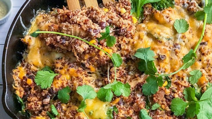 A skillet brimming with a vibrant chicken quinoa bake, crowned with melted cheese, fresh spinach, and cilantro. A wooden spatula rests in the flavorful dish, while plates are stacked nearby in anticipation.