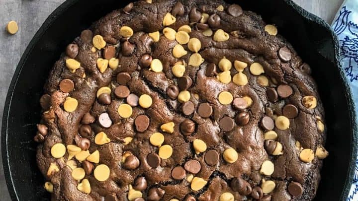 A delicious skillet cookie topped with chocolate and peanut butter chips sits on a wooden surface beside a wooden spoon, a plate with forks, and a blue-patterned napkin.