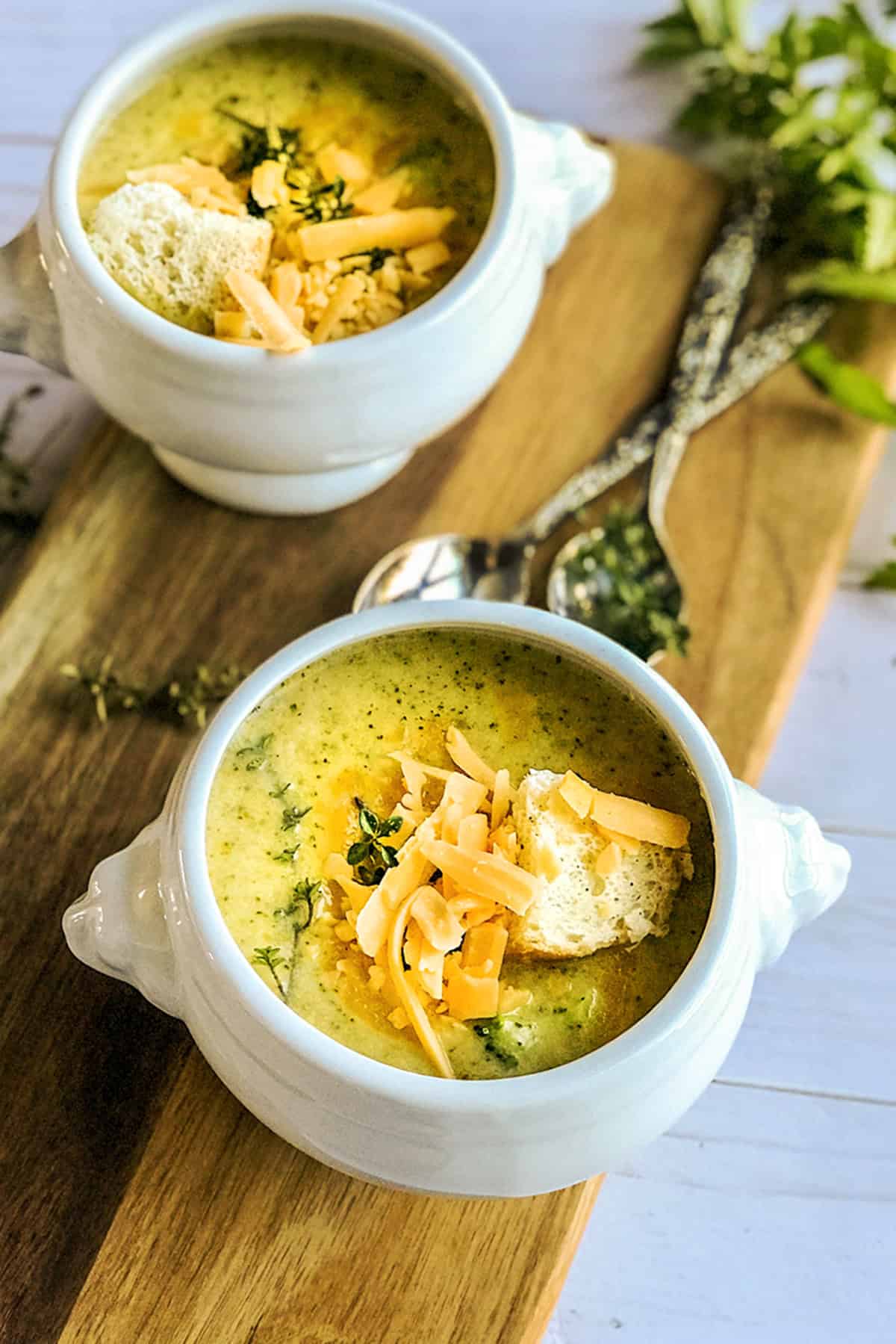 Two bowls of broccoli cheddar soup in footed bowls and topped with fresh thyme, shredded cheddar and bread squares.