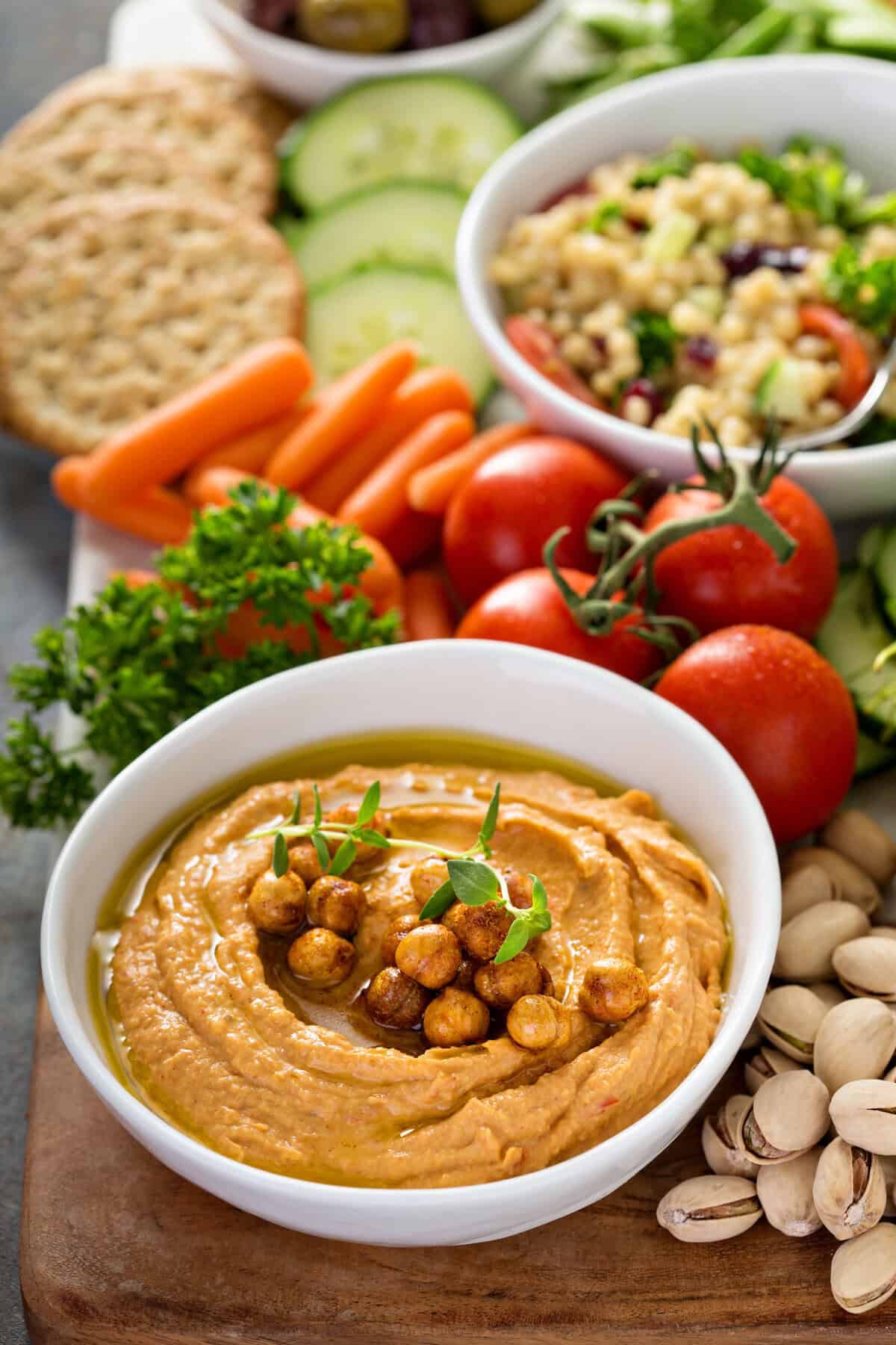 A bowl of red pepper hummus topped with roasted chickpeas, surrounded by fresh vegetables, tomatoes, crackers, pistachios, and a bowl of salad on a wooden board.