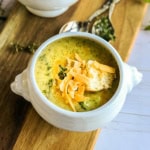 Bowl of broccoli cheddar soup in a footed bowl on a wooden serving board with shredded cheese, herbs, and bread cubes as garnish.