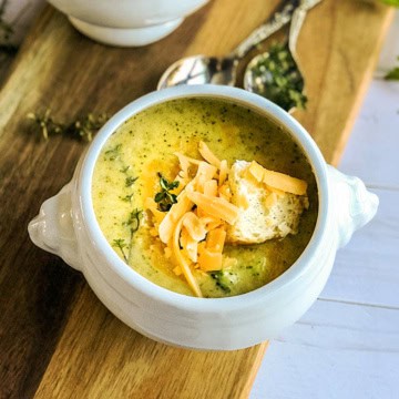 Bowl of broccoli cheddar soup in a footed bowl on a wooden serving board with shredded cheese, herbs, and bread cubes as garnish.