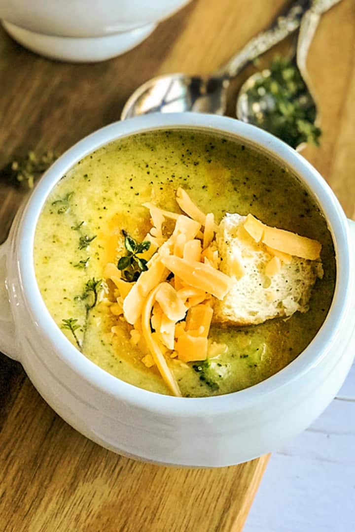 Bowl of broccoli cheddar soup in a footed bowl on a wooden serving board with shredded cheese, herbs, and bread cubes as garnish.