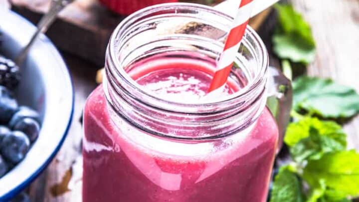 A pink triple berry smoothie in a mason jar with a red and white striped straw, surrounded by fresh strawberries, blueberries, and mint on a wooden table.