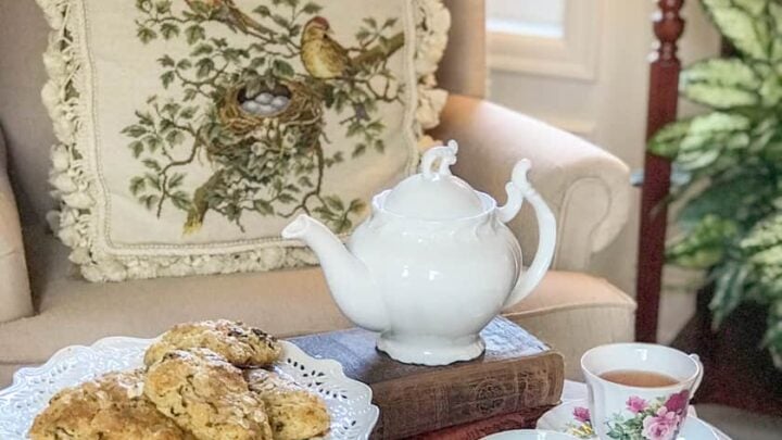 A white teapot, a plate of scones, and two teacups sit on a small table next to an armchair with a bird-themed pillow and plaid blanket, creating the perfect setting for a cozy Winter Afternoon Tea.