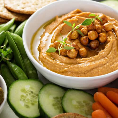 A bowl of red pepper hummus topped with chickpeas and herbs, surrounded by sliced cucumbers, cherry tomatoes, carrots, snap peas, crackers, and a bowl of olives.