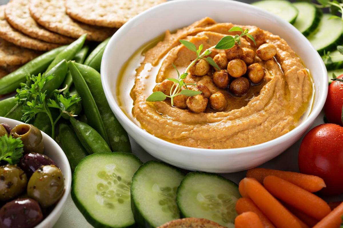 A bowl of red pepper hummus topped with chickpeas and herbs, surrounded by sliced cucumbers, cherry tomatoes, carrots, snap peas, crackers, and a bowl of olives.