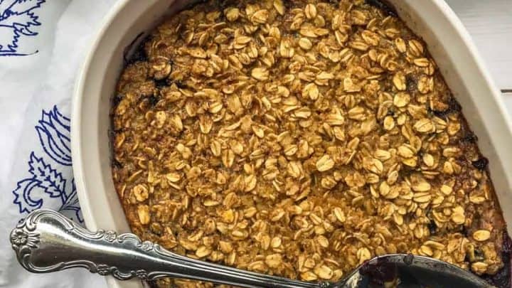 A white baking dish with blueberry amish baked oatmeal and berries, partially served with a large spoon on a white table next to a cup of coffee and stacked plates.