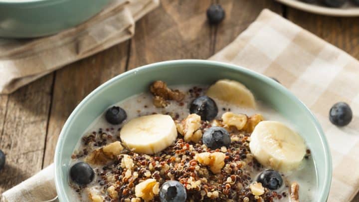 A bowl of breakfast quinoa cereal topped with banana slices, blueberries, and walnuts, with milk, sits on a wooden table next to a plate of fresh blueberries.