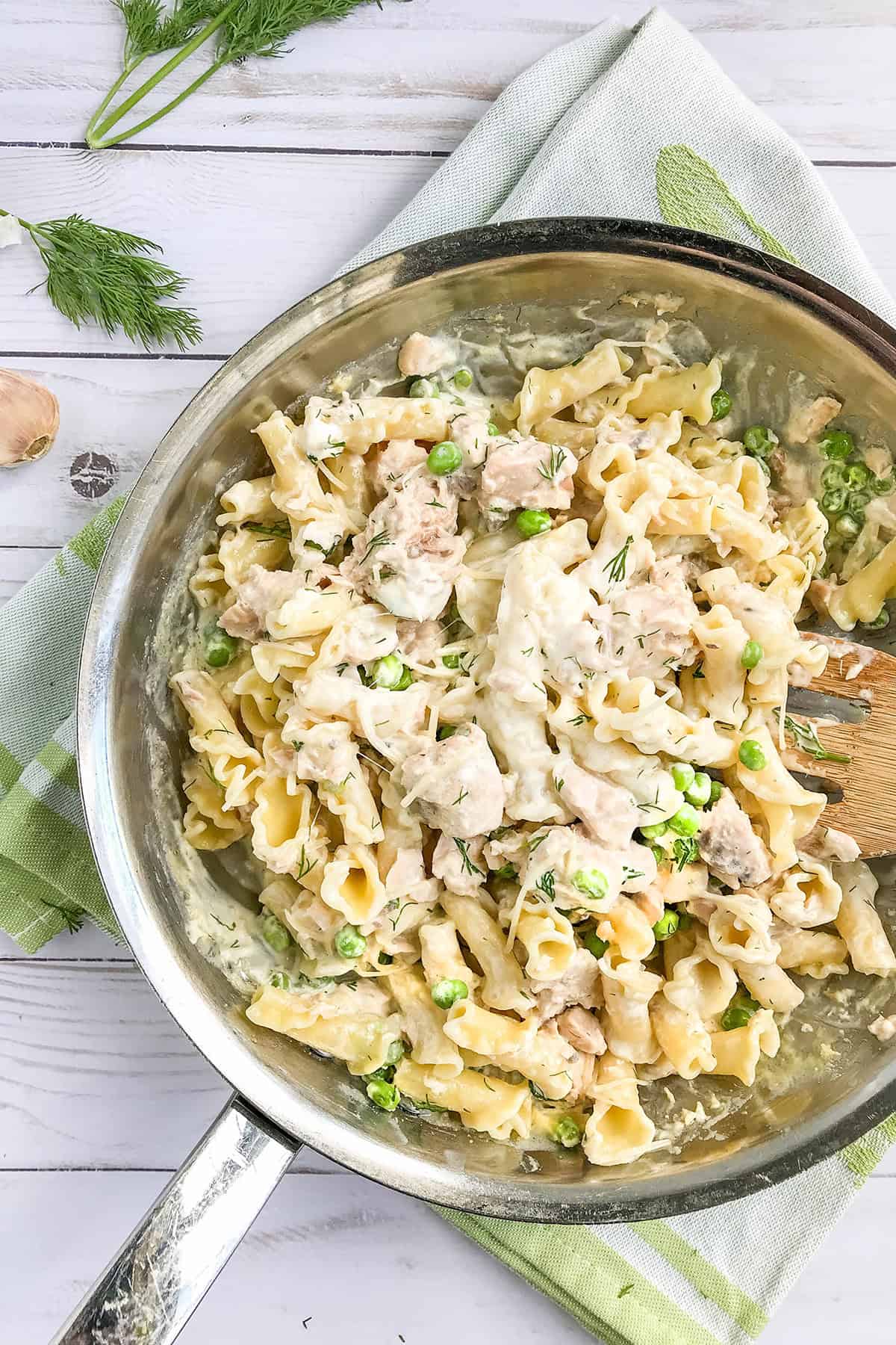 A skillet filled with salmon pasta, tender chicken pieces, green peas, and a creamy sauce, garnished with fresh dill on a white wooden surface.