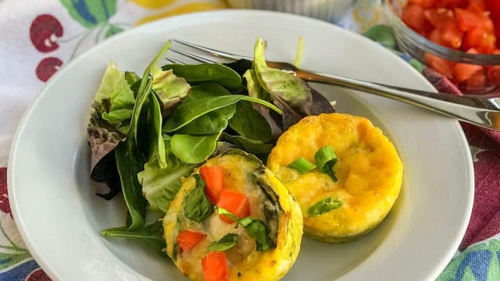 A white plate with two mini frittatas topped with diced red peppers and a side of mixed greens; small bowls of chopped green onions and tomatoes in the background.
