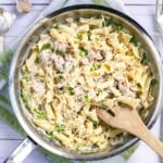 A skillet of creamy salmon pasta with shredded chicken, green peas, fresh herbs, and grated parmesan, with a wooden spoon resting inside.
