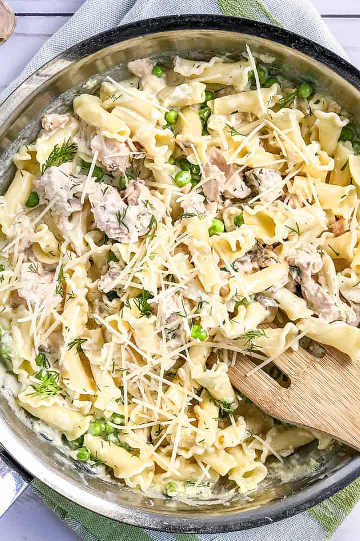 A skillet of creamy salmon pasta with shredded chicken, green peas, fresh herbs, and grated parmesan, with a wooden spoon resting inside.