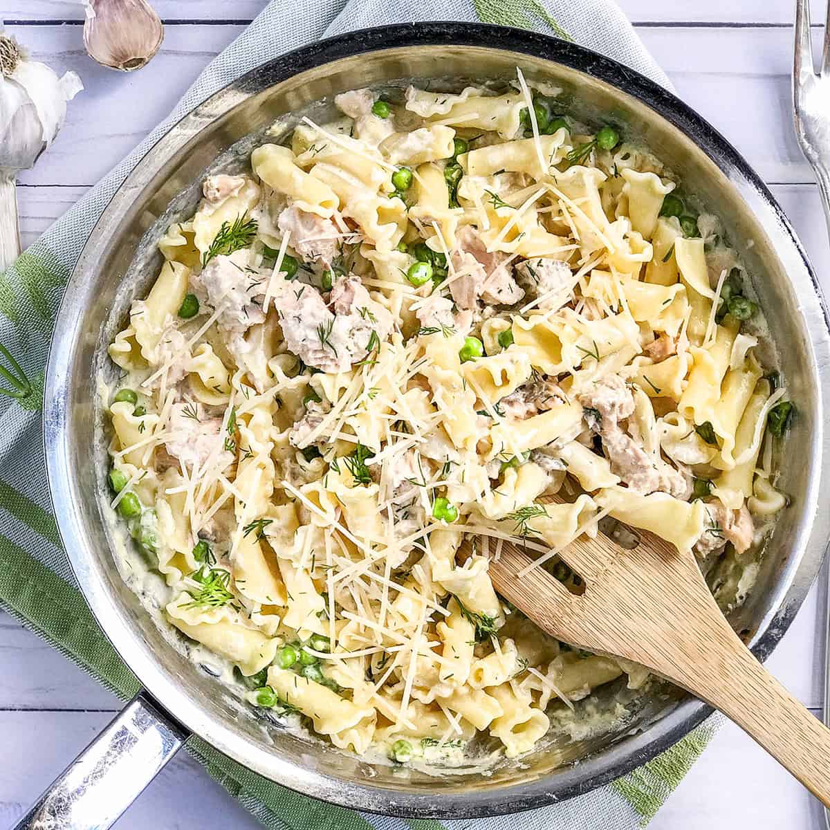 A skillet of creamy salmon pasta with shredded chicken, green peas, fresh herbs, and grated parmesan, with a wooden spoon resting inside.