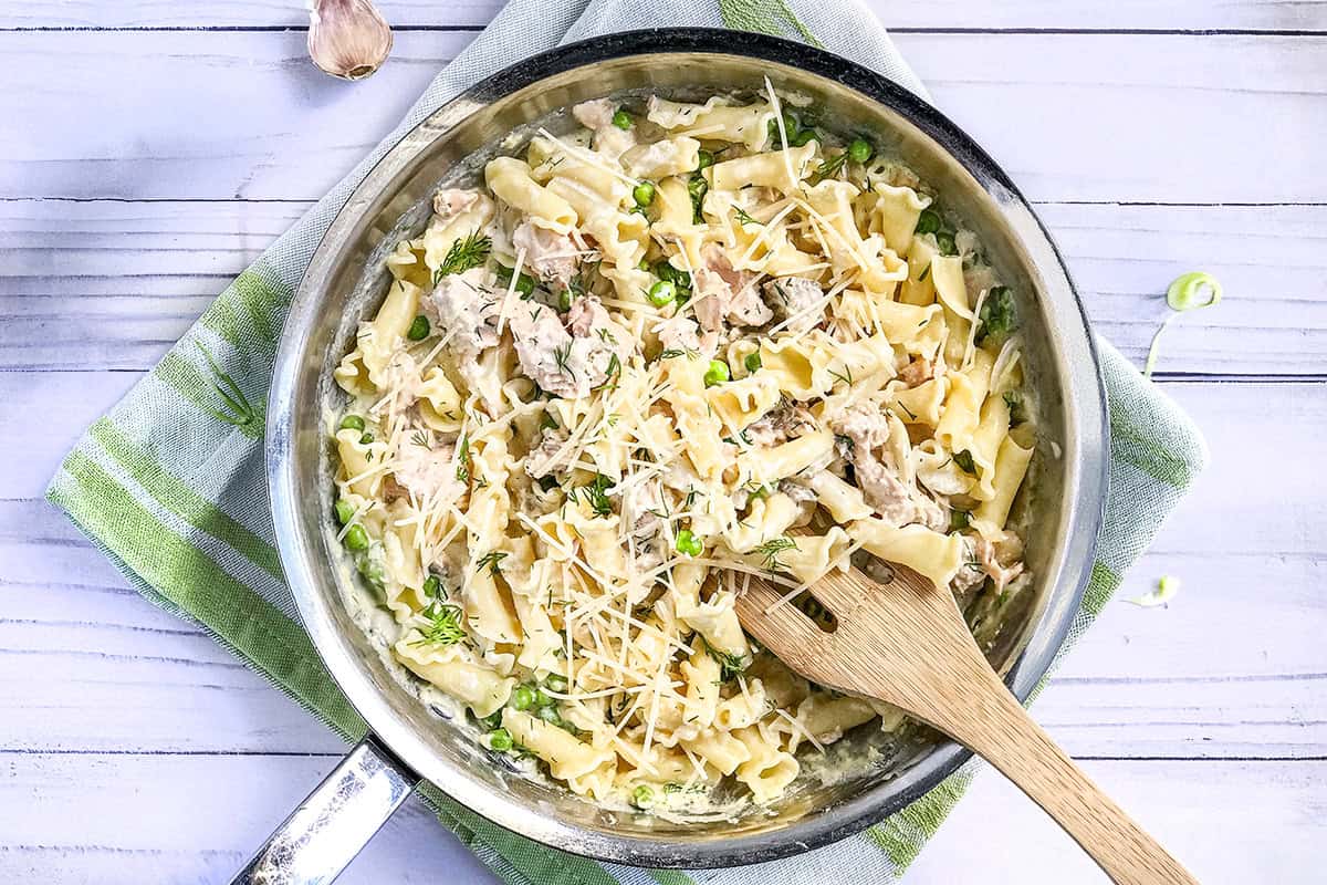 A skillet of salmon pasta with green peas, herbs, and grated cheese, stirred with a wooden spoon, sits on a green and white towel.