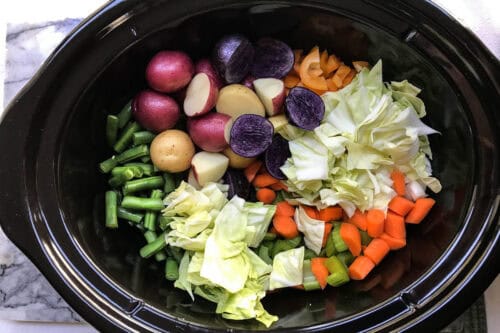 Top view of a slow cooker filled with vibrant ingredients for a healthy cabbage soup, featuring assorted vegetables like red and purple potatoes, chopped cabbage, sliced carrots, green beans, and orange bell peppers, all arranged in sections before cooking.