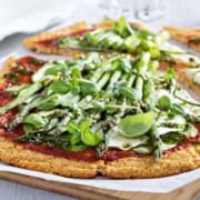 A cauliflower pizza with a thin crust topped with tomato sauce, zucchini slices, asparagus, and fresh greens, served on parchment paper.